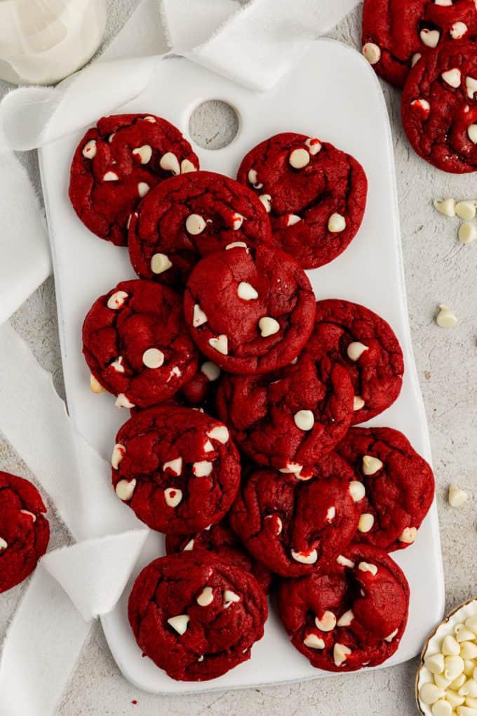 Biscotti Red Velvet con preparato per torte su un tagliere bianco circondato da gocce di cioccolato bianco sparse e altri biscotti