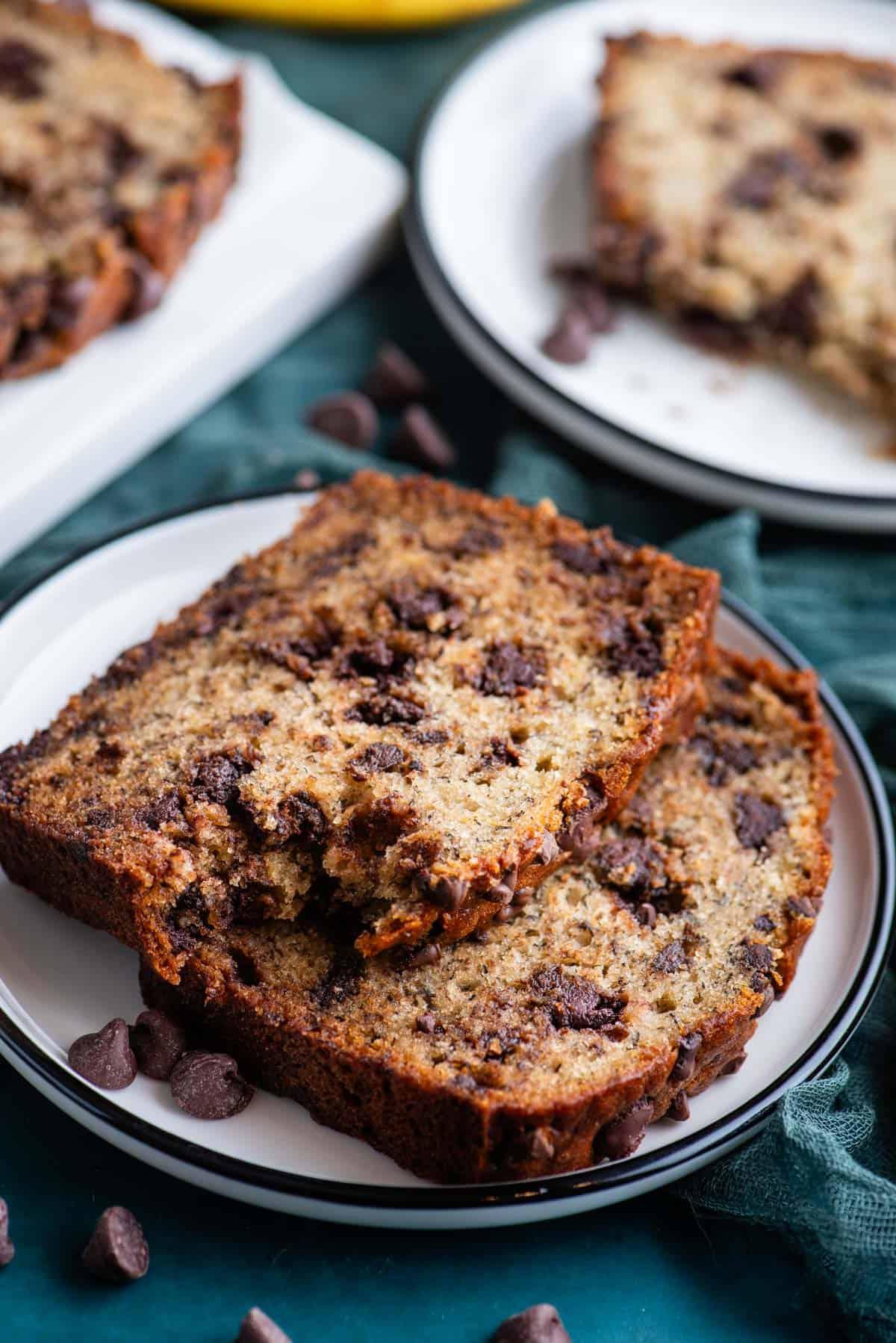 Due fette di pane alle banane e gocce di cioccolato su un piccolo piatto bianco su un asciugamano color ottanio scuro, con un morso preso dalla fetta superiore, un altro piatto con pane e un tagliere bianco con pane sullo sfondo e gocce di cioccolato sparse intorno.