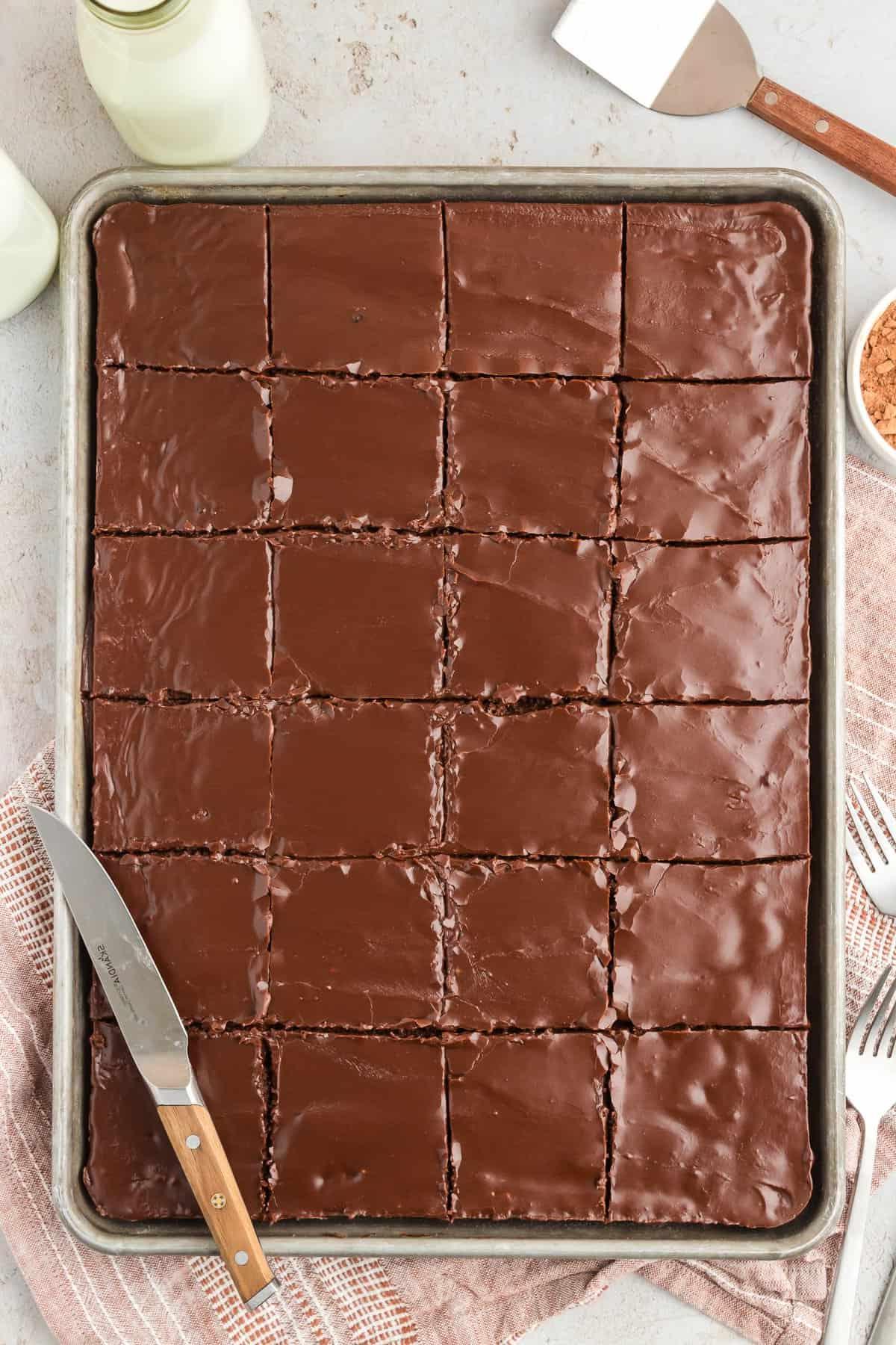 Una teglia di torta Texana tagliata a quadrati con un coltello appoggiato sopra, una spatola, latte, una ciotola di cacao in polvere, una forchetta e un panno da cucina color cuoio intorno