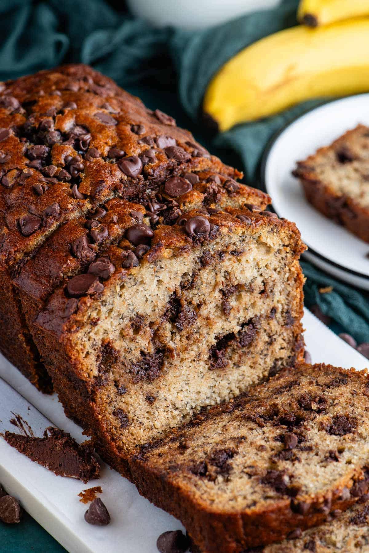 Pane alle banane e gocce di cioccolato affettato su un tagliere bianco con un piatto sullo sfondo con una fetta e banane intere dietro.