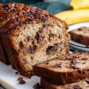 Pane alle banane e gocce di cioccolato affettato su un tagliere bianco con un piatto sullo sfondo con una fetta e banane intere dietro.