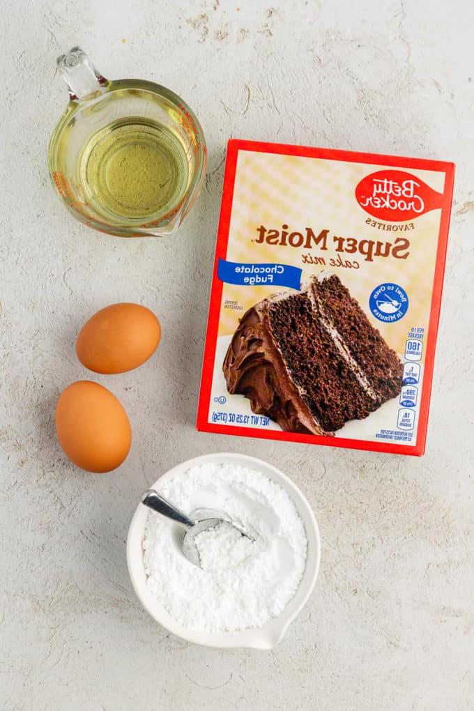 Ingredienti per biscotti Chocolate Crinkle con preparato per torte, inclusi una scatola di preparato per torte al cioccolato, olio vegetale, due uova e una piccola ciotola bianca di zucchero a velo