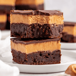 Due brownies decadenti impilati su un piatto bianco, ciascuno con una base di cioccolato fondente, uno spesso strato cremoso di burro d'arachidi e una lucida copertura di cioccolato. Altri brownies sono sfocati sullo sfondo.