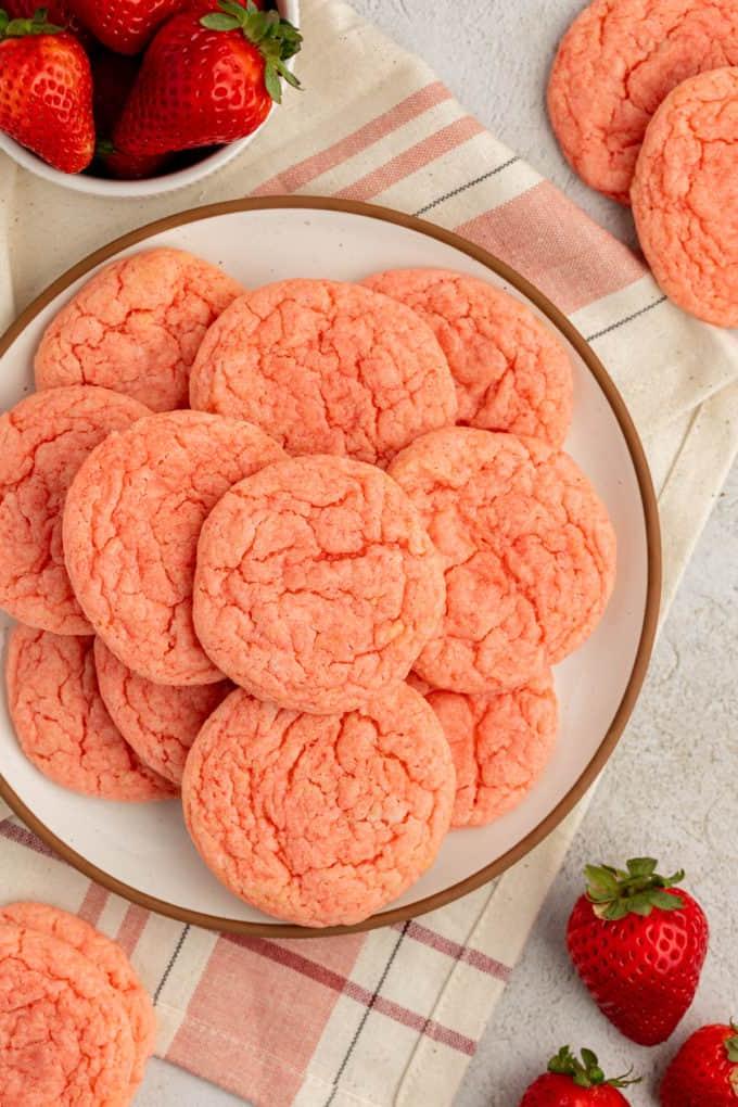Biscotti alla fragola con preparato per torte impilati su un piatto bianco con bordo marrone, appoggiato su un canovaccio da cucina beige e rosa e circondato da altri biscotti, fragole e una piccola ciotola bianca di fragole