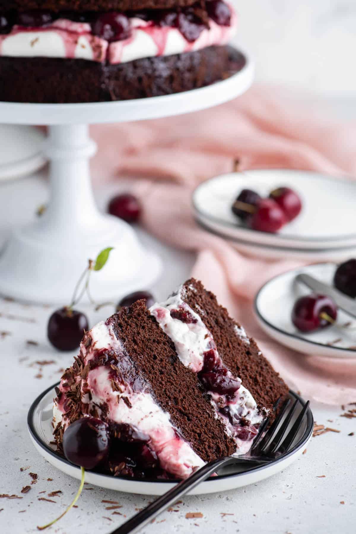 Una fetta di Torta Foresta Nera su un piatto bianco con un'alzatina e altri piatti sullo sfondo e ciliegie e scaglie di cioccolato sparse intorno