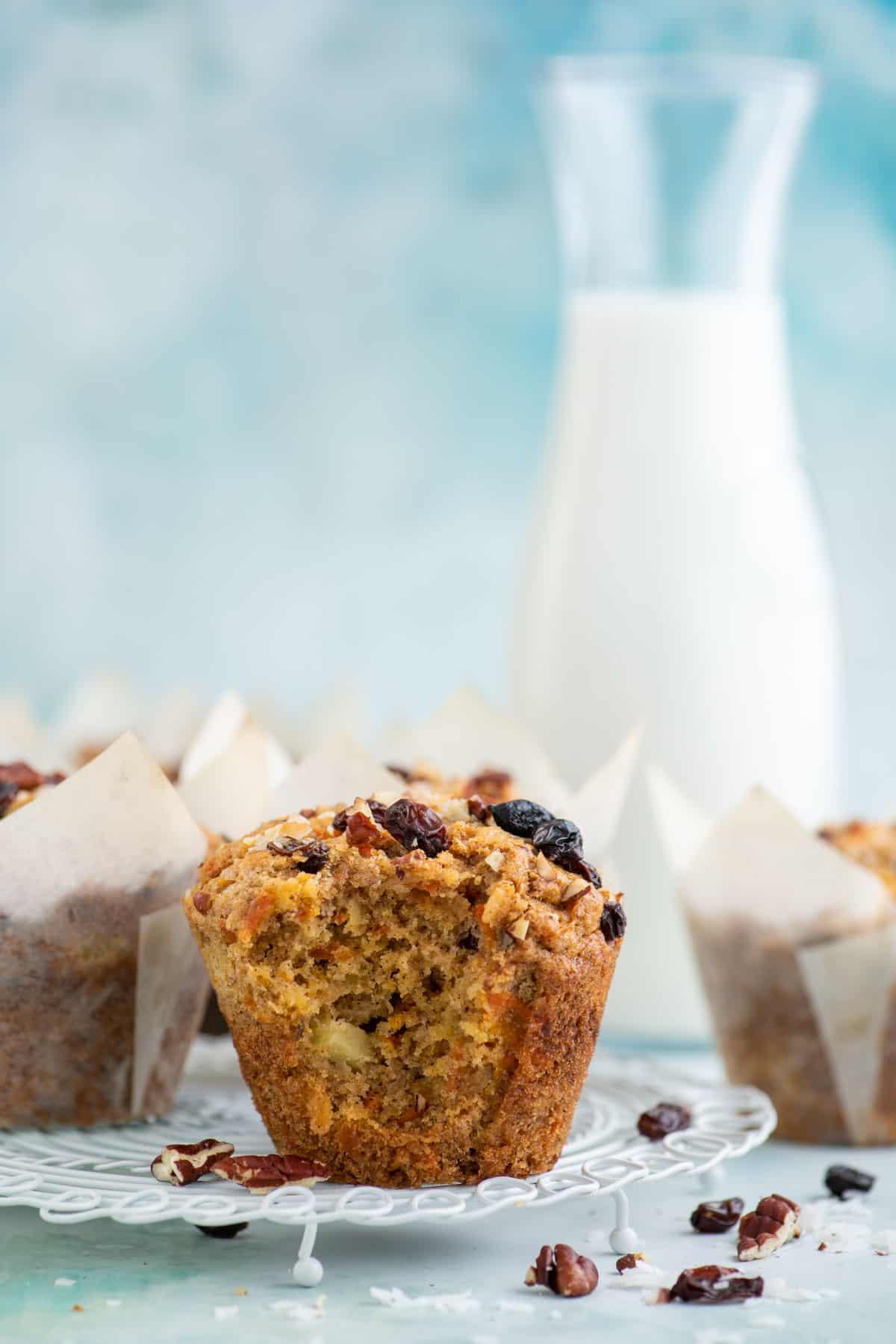Un muffin Morning Glory parzialmente mangiato su una griglia bianca, circondato da altri muffin avvolti nella carta, noci, uvetta e scaglie di cocco.