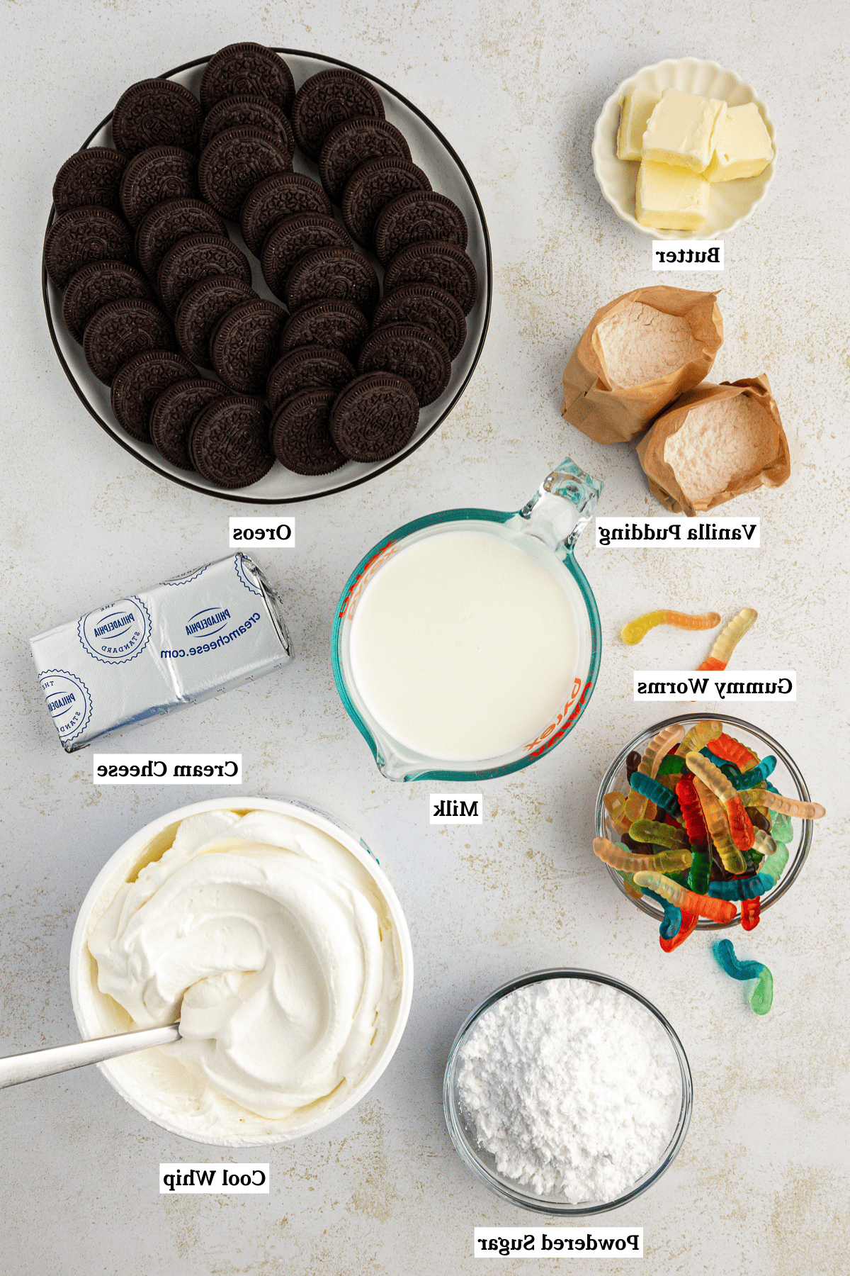 Vista dall'alto degli ingredienti per la torta di terra, inclusi burro, preparato per budino istantaneo alla vaniglia, latte, Oreo, vermi gommosi, crema di formaggio, Cool Whip e zucchero a velo