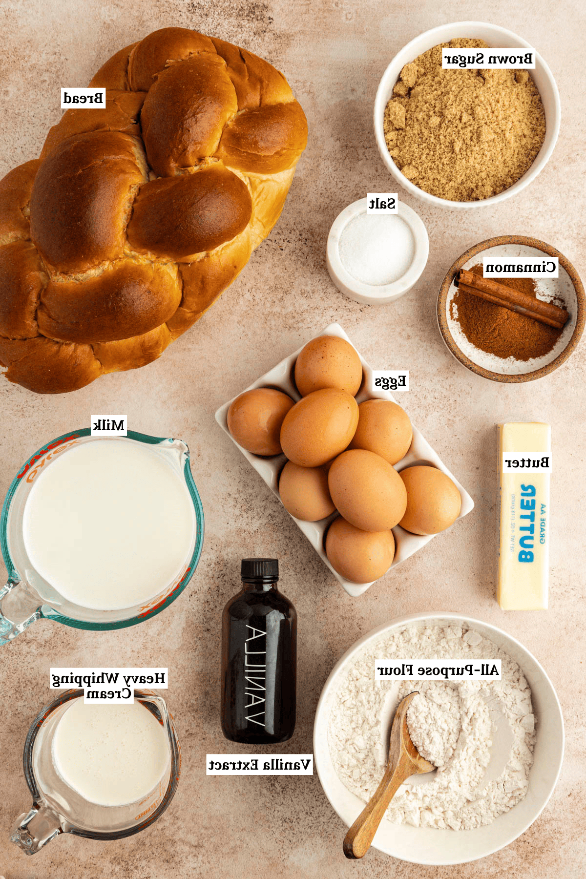 Vista dall'alto degli ingredienti per la casseruola di French Toast, inclusi una pagnotta di pane, una ciotola di zucchero di canna, una piccola ciotola di sale, una ciotola di cannella, otto uova marroni, un panetto di burro, una tazza di latte, una tazza di panna da montare, una bottiglia di estratto di vaniglia e una ciotola di farina per tutti gli usi.