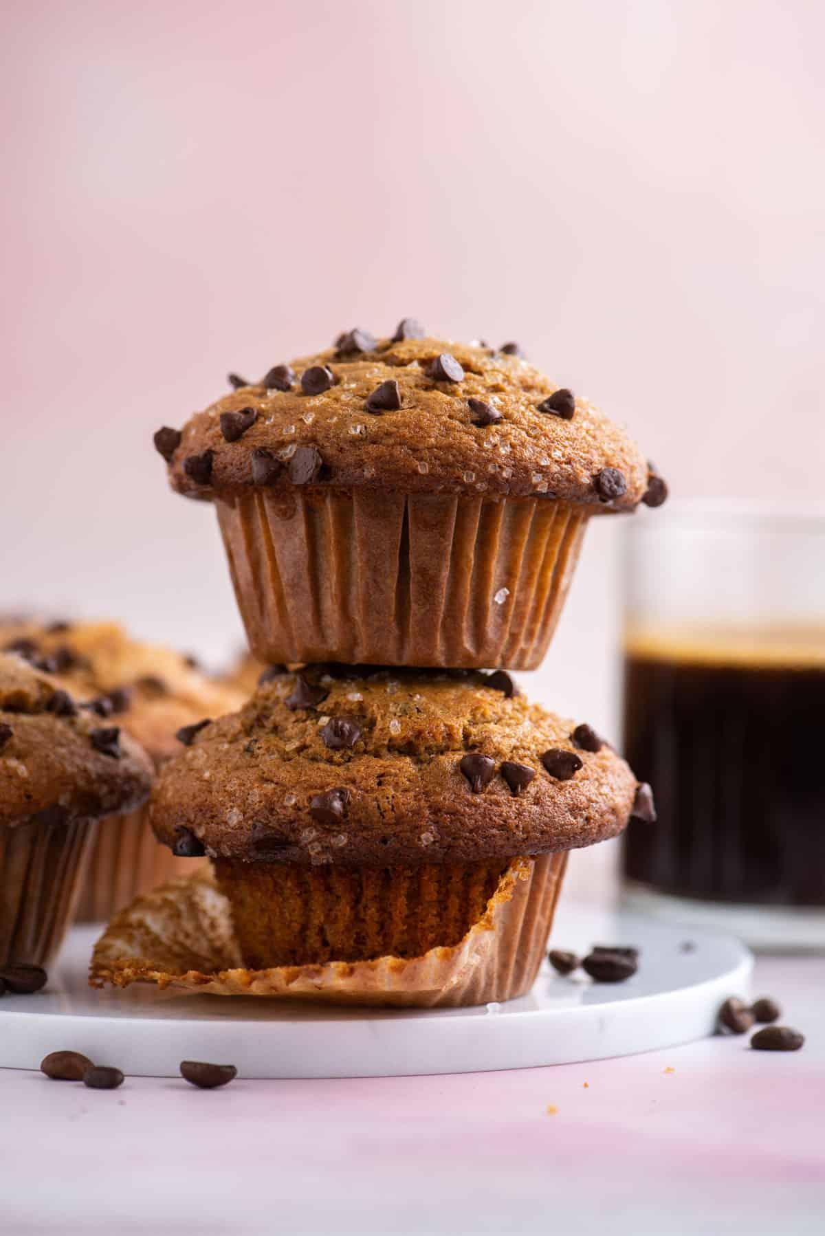 Muffin al Cappuccino e Gocce 4 una pila di due muffin al cappuccino con gocce di cioccolato con chicchi di caffè sparsi intorno, altri muffin e una tazza di caffè sullo sfondo