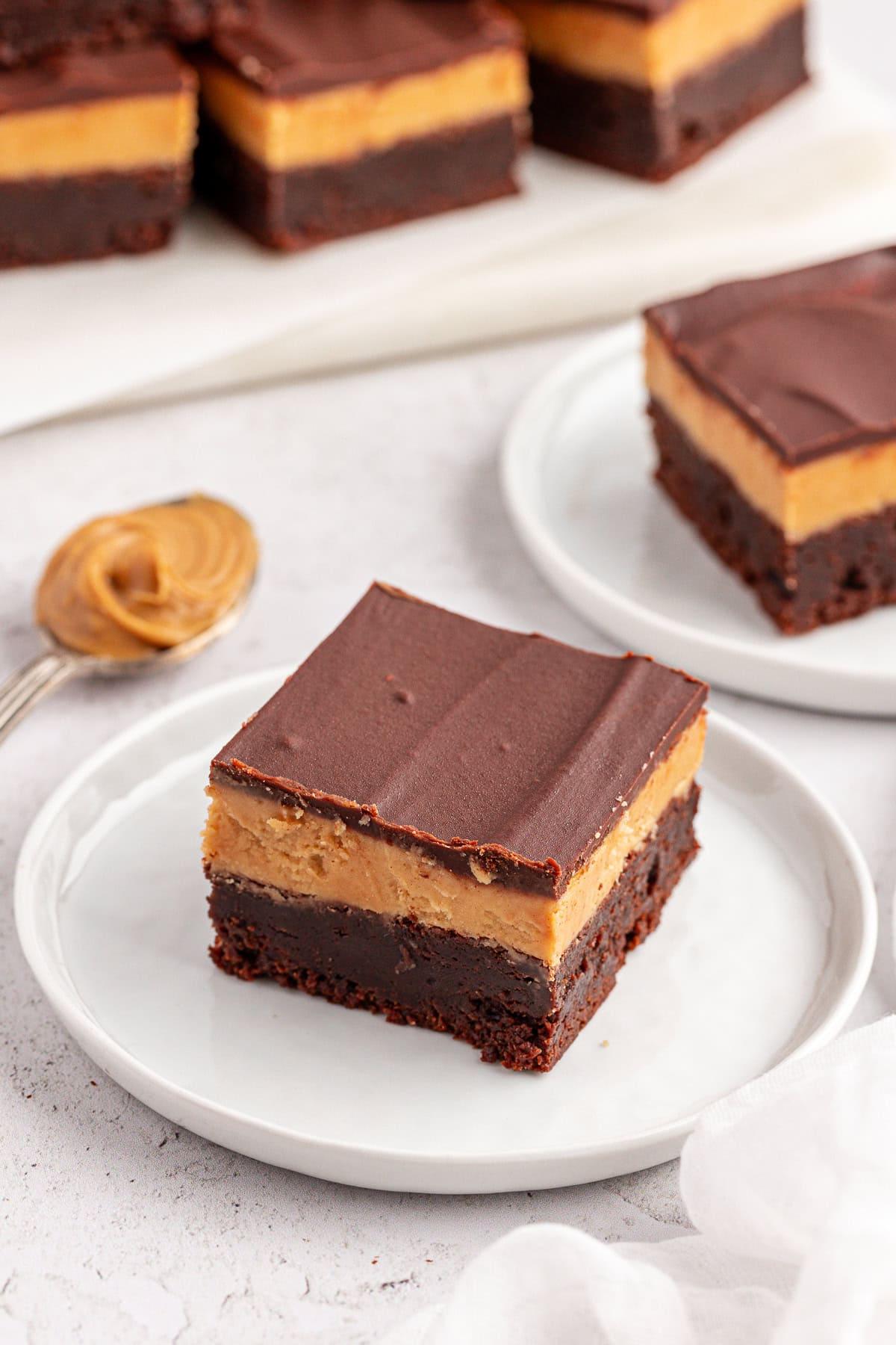 Primo piano di un brownie Buckeye a tre strati: una base densa di brownie, un ripieno cremoso al burro d'arachidi e una morbida copertura di cioccolato, servito su un piatto bianco. Un cucchiaio con burro d'arachidi e altre barrette di dessert sono sullo sfondo.