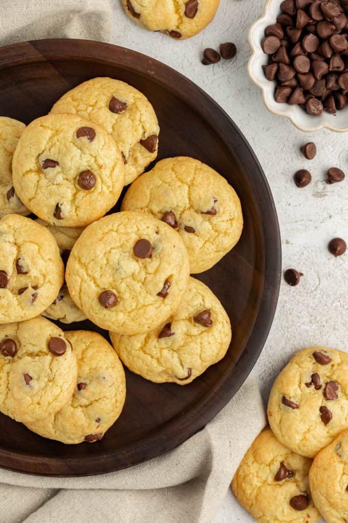 Biscotti con gocce di cioccolato e preparato per torte su un piatto marrone su un canovaccio di lino beige, circondati da gocce di cioccolato sparse, altri biscotti e una piccola ciotola bianca di gocce di cioccolato