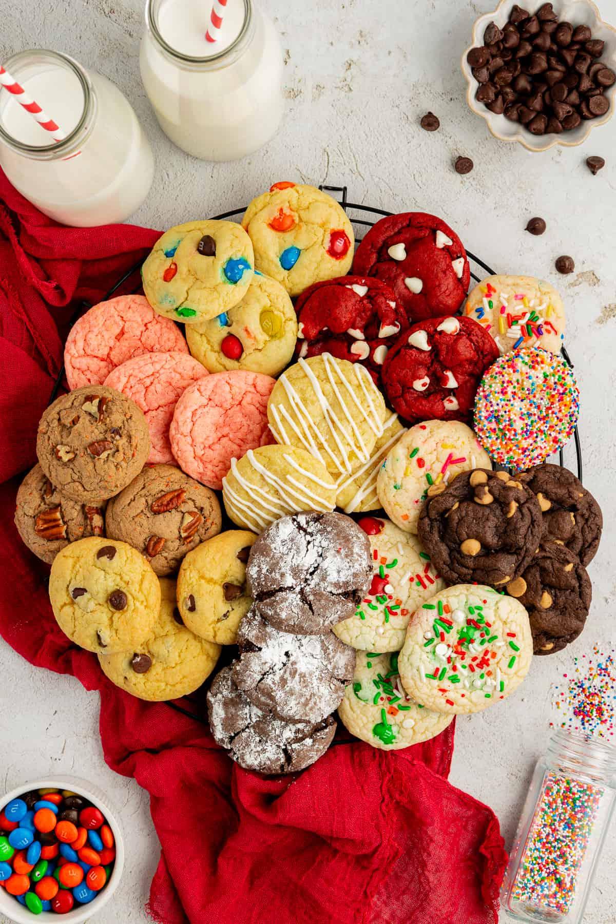 Un assortimento di biscotti con preparato per torte di diversi gusti in cerchio su una griglia su un canovaccio rosso con codette, M&M's, gocce di cioccolato e due bicchieri di latte con cannucce rosse e bianche intorno