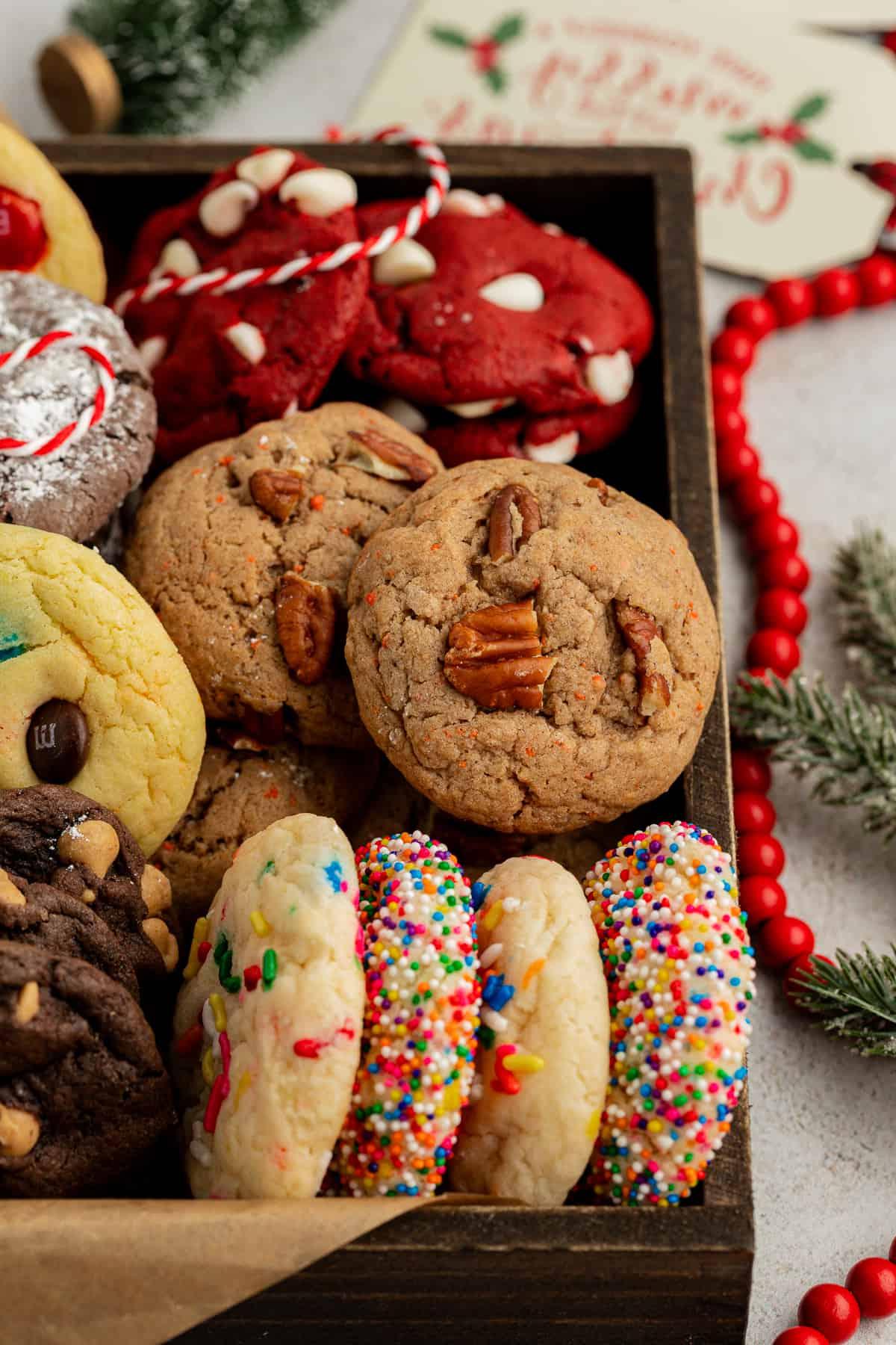 Assortimento di biscotti con preparato per torte di diversi gusti in una scatola di legno con carta marrone circondata da perline rosse, rami di pino e spago rosso e bianco