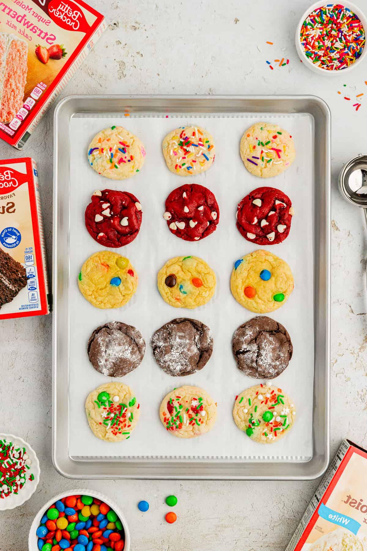 Assortimento di biscotti con preparato per torte di diversi gusti in file su una teglia rivestita di carta forno circondata da scatole di preparato per torte, codette colorate, M&M's e uno scoop per biscotti