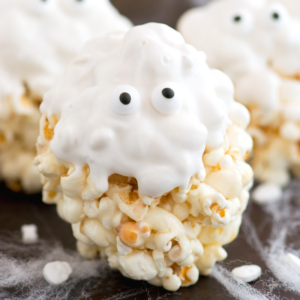 Una palla di popcorn fantasma ricoperta di glassa bianca e decorata con due occhietti di zucchero, che assomiglia a una spettrale palla di popcorn di Halloween. Il dolcetto è posato su una superficie nera con sottili filamenti bianchi che ricordano ragnatele.