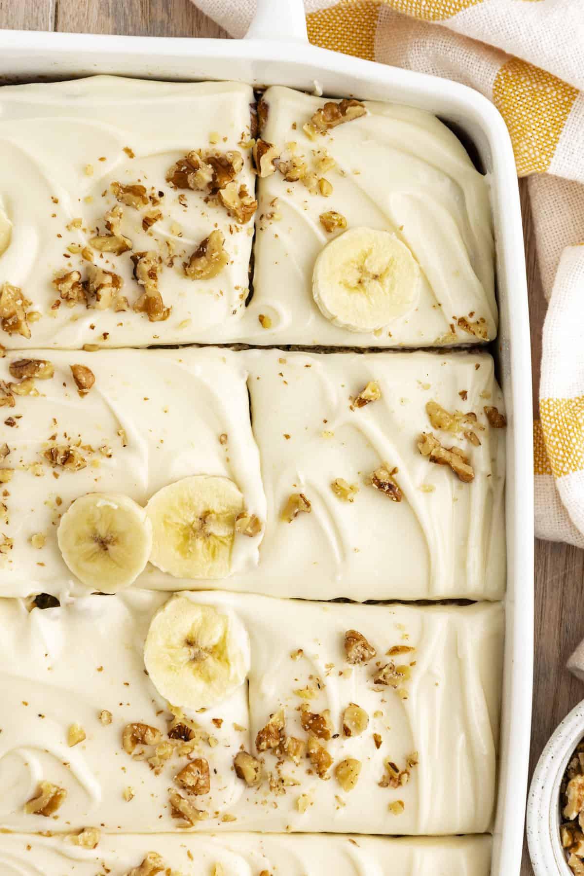 Vista dall'alto di una torta di banane in una teglia bianca, tagliata a pezzi e guarnita con fette di banana e noci tritate, con un canovaccio da cucina a strisce gialle e bianche accanto alla teglia