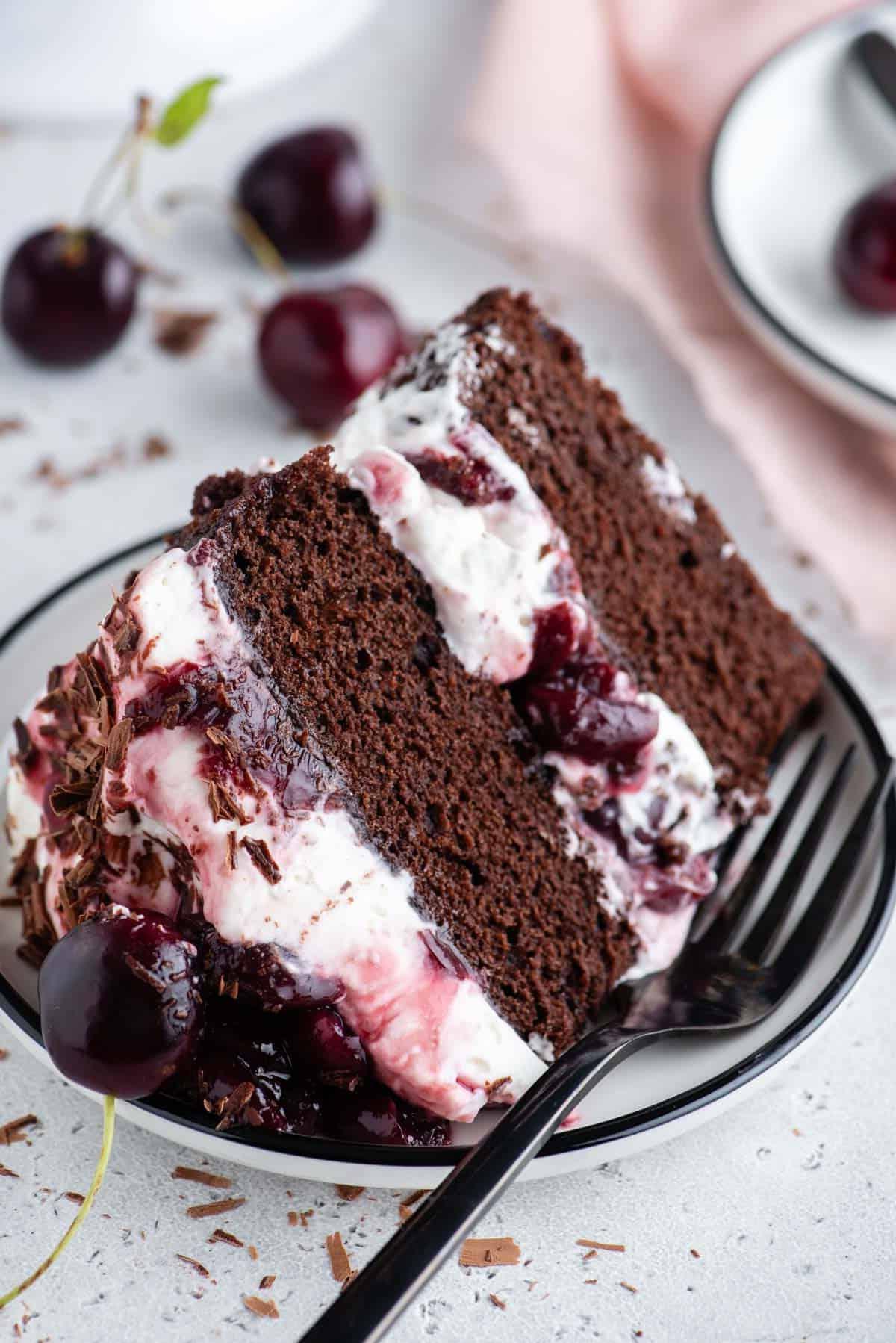 Una fetta di torta Foresta Nera su un piatto con una forchetta, circondata da ciliegie e piccoli pezzi di cioccolato a scaglie