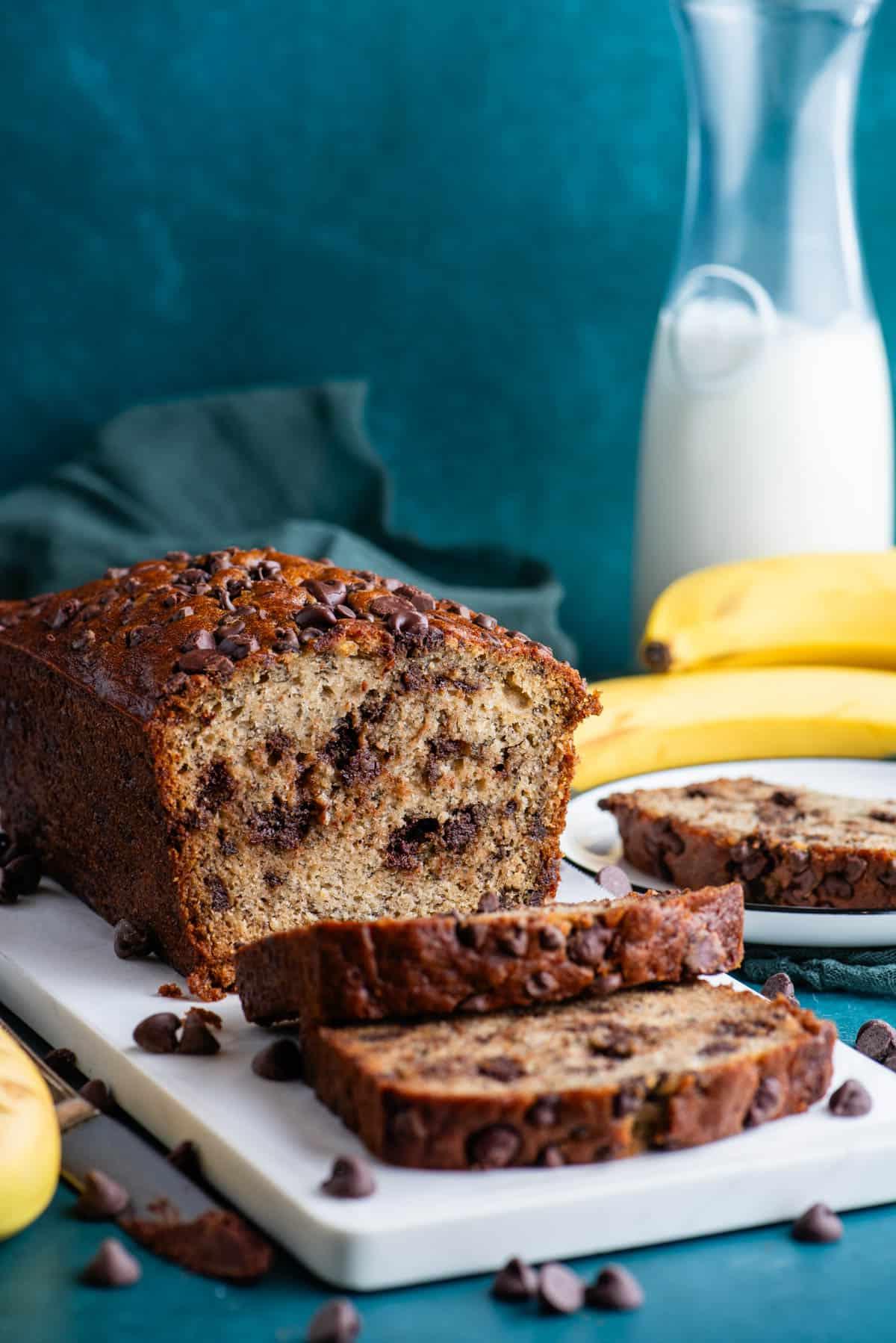 Una pagnotta di pane alle banane e gocce di cioccolato su un tagliere bianco con un piatto con una fetta di pane alle banane accanto, gocce di cioccolato sparse intorno, e banane intere e un bicchiere di latte sullo sfondo.