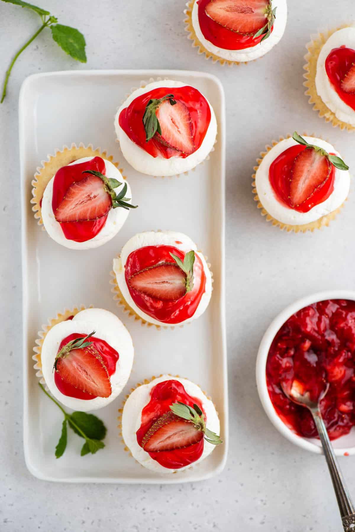 I Cupcake alle Fragole Shortcake sono fatti con torta alla vaniglia umida, ripieni di dolce salsa di fragole e ricoperti di soffice panna montata! Cupcake alle fragole shortcake disposti su un piccolo piatto bianco