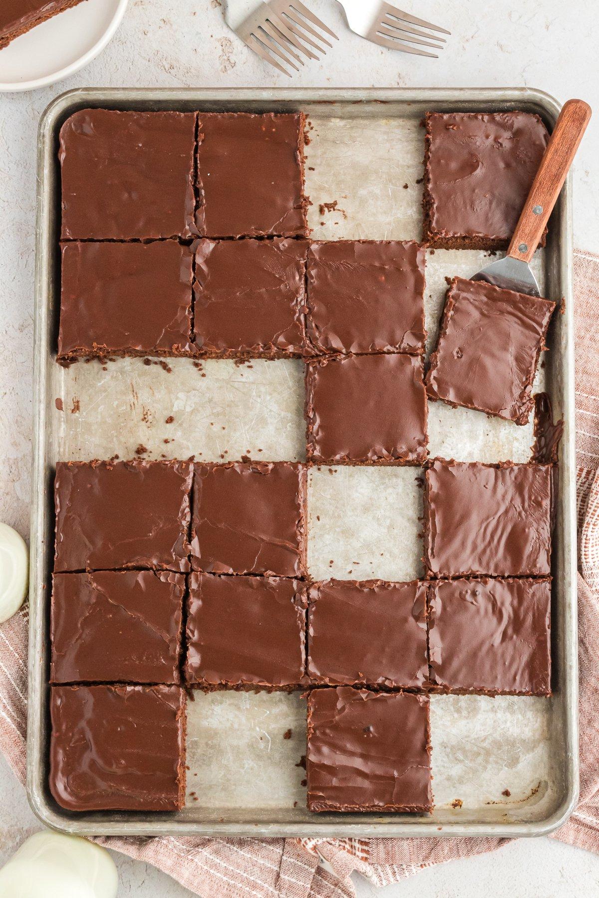 Fette quadrate di torta Texana su una grande teglia con alcune fette mancanti, una spatola sotto una fetta, alcune forchette e un piatto con una fetta di torta accanto