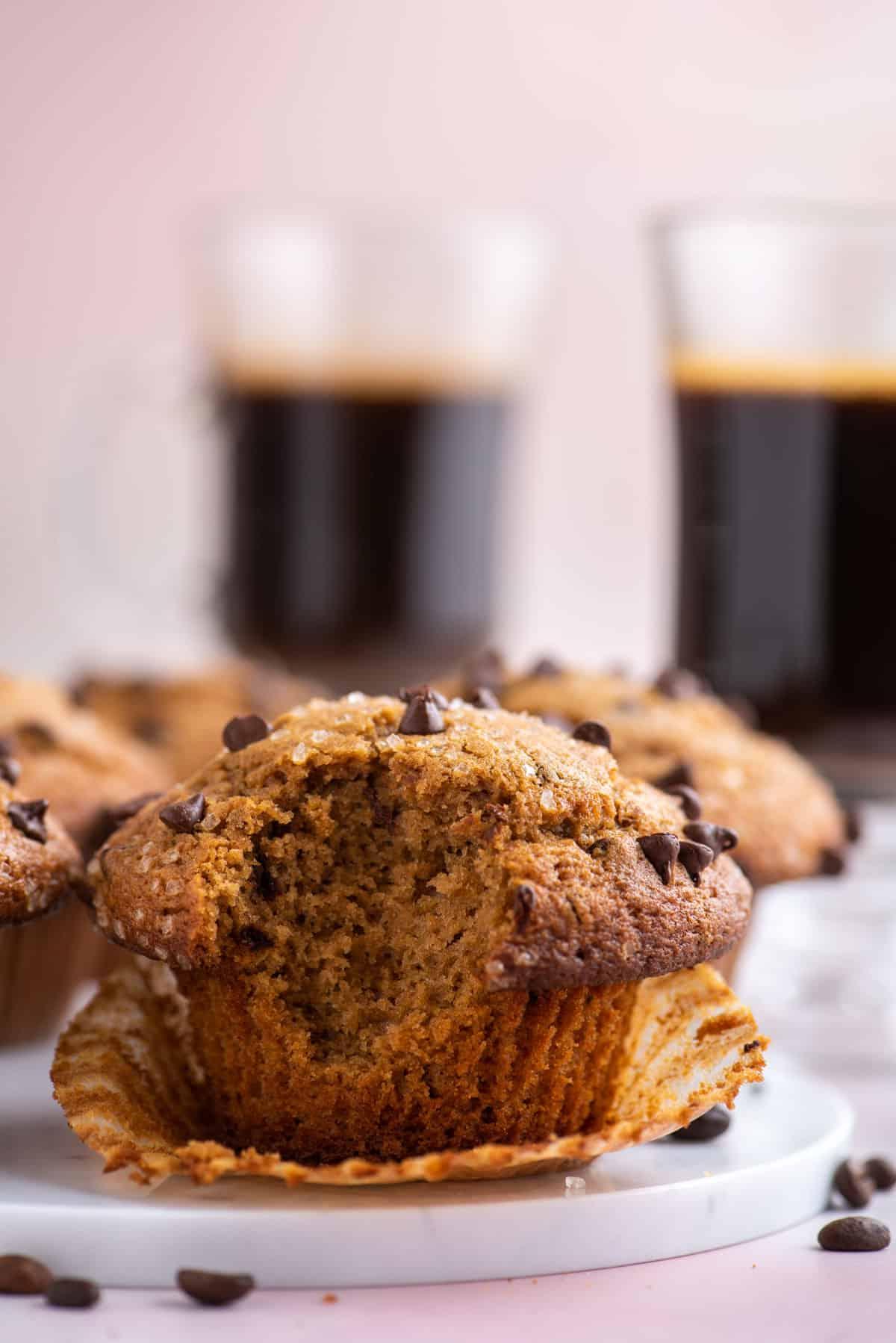Muffin al Cappuccino e Gocce 5 un muffin al cappuccino con gocce di cioccolato su un piatto bianco con un morso preso, appoggiato nel suo pirottino aperto, con chicchi di caffè sparsi intorno, altri muffin e due tazze di caffè sullo sfondo