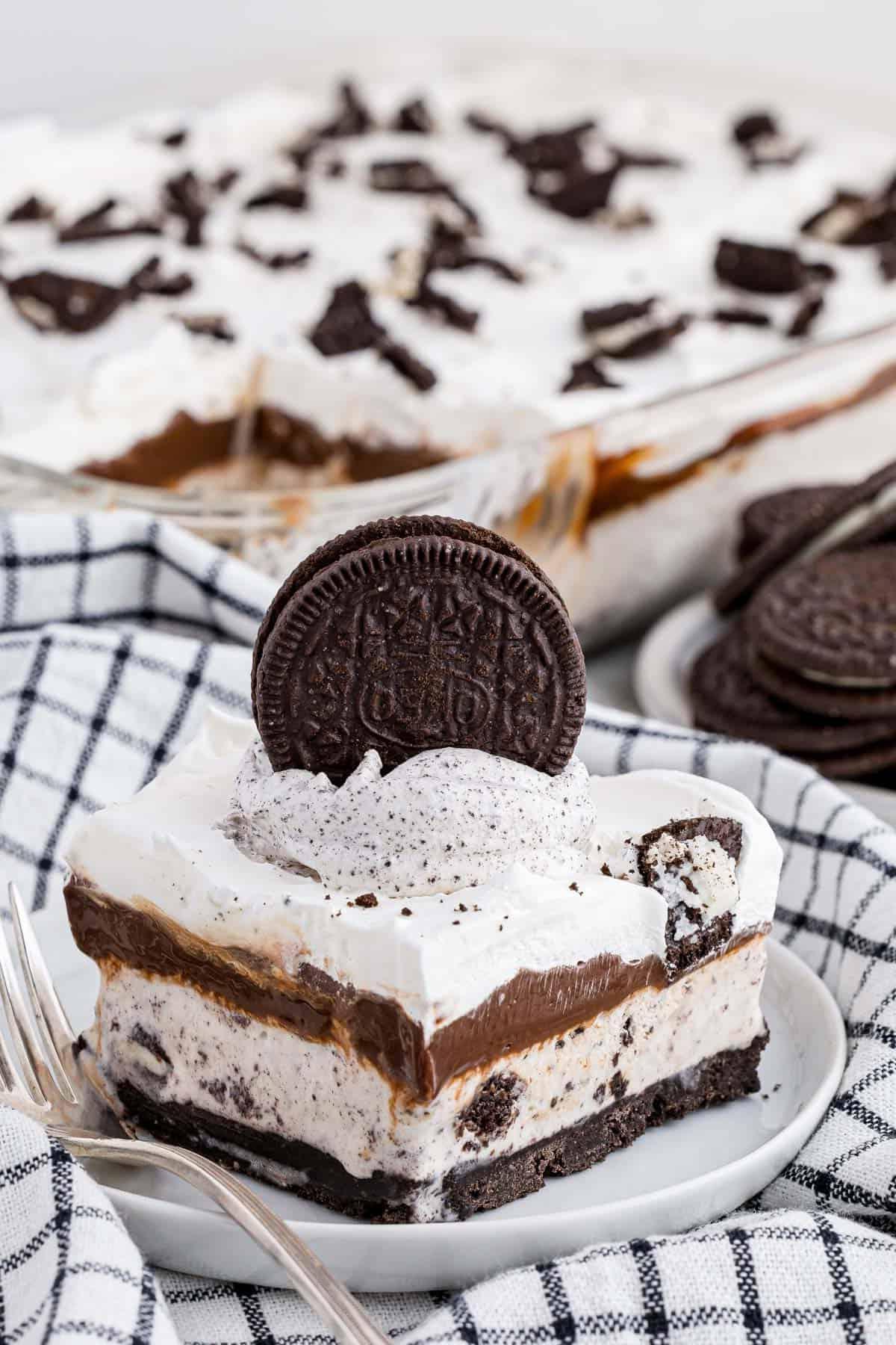 Ricetta Torta Gelato Oreo con 5 Ingredienti 5 Una fetta di torta gelato Oreo su un piatto bianco con una forchetta, poggiata su un canovaccio da cucina a scacchi bianchi e neri con altra torta gelato e Oreo sullo sfondo