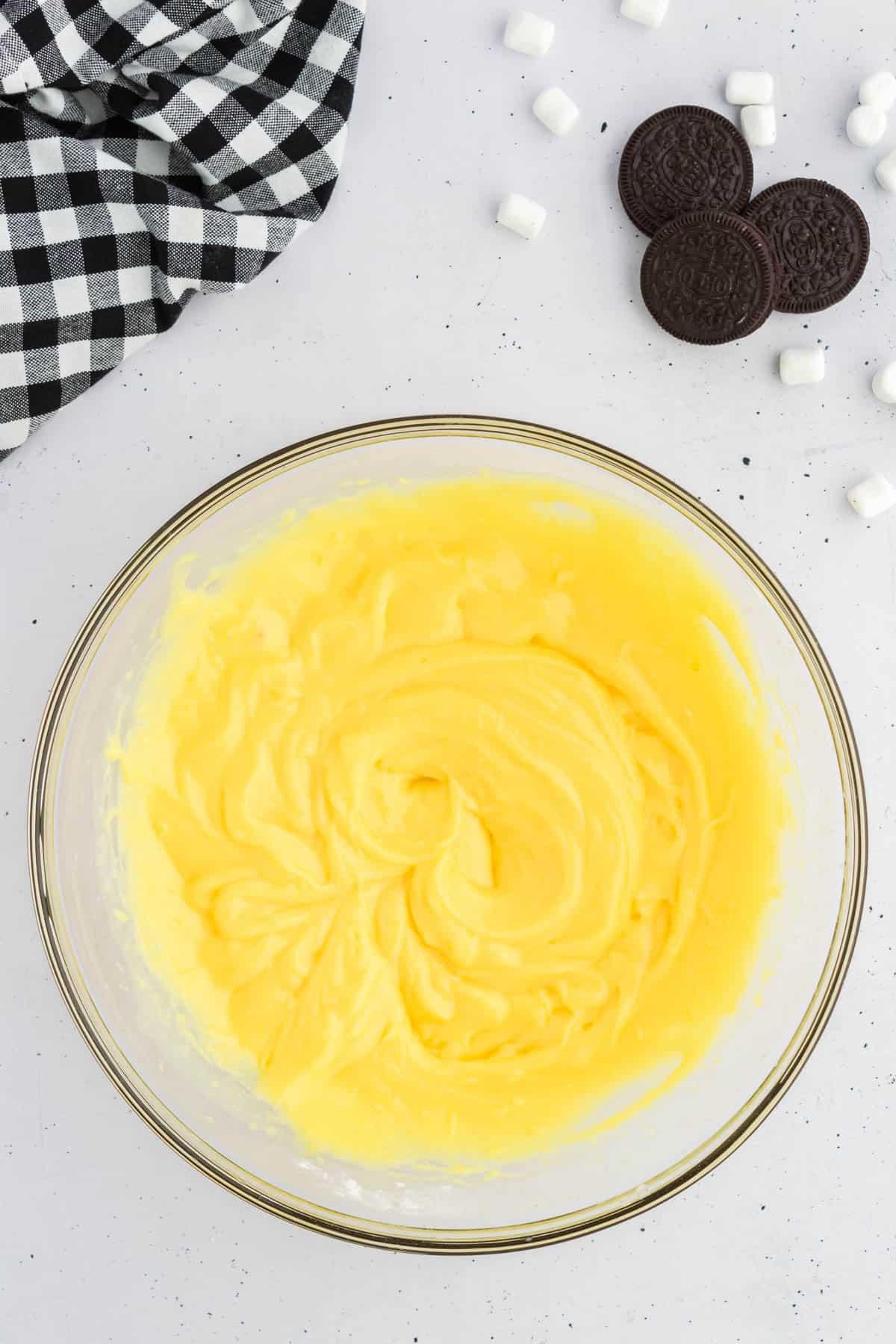 Budino alla vaniglia in una ciotola di vetro circondato da Oreo, mini marshmallow e un panno da cucina a scacchi bianchi e neri
