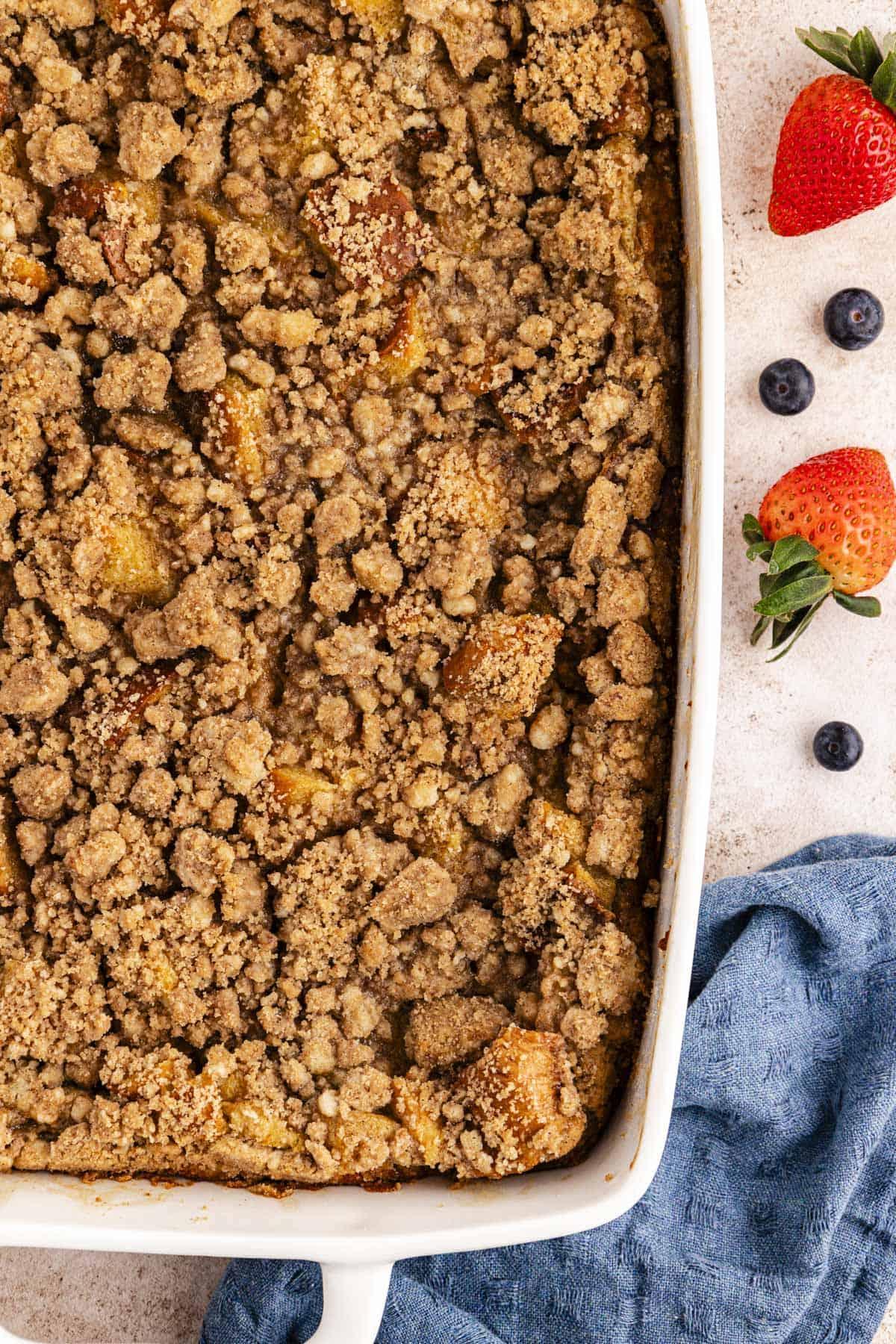 Una grande teglia bianca di casseruola di French Toast con topping croccante accanto a un canovaccio blu e mirtilli e fragole fresche sparse intorno.