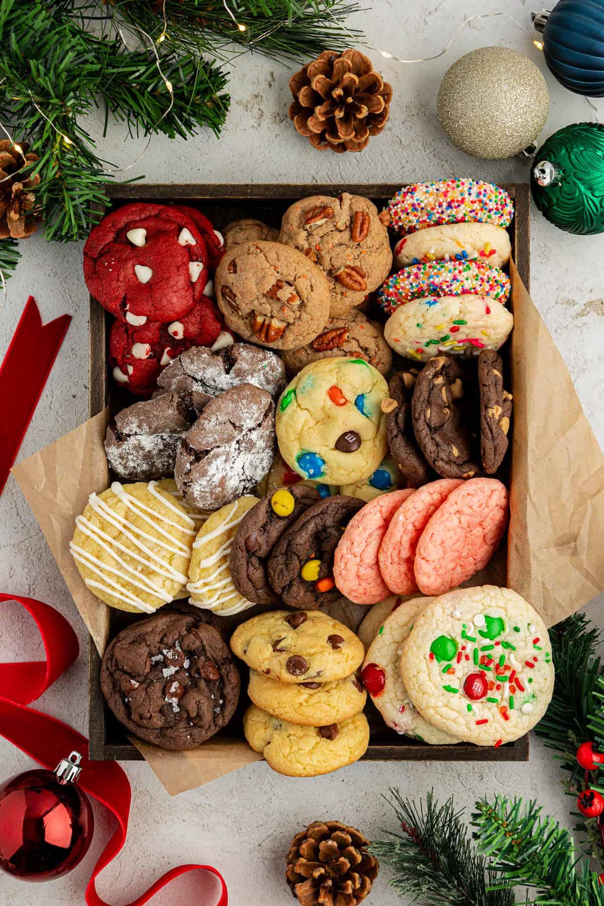 Una scatola piena di biscotti con preparato per torte di vari gusti con carta marrone circondata da ornamenti natalizi, nastro rosso, pigne e rami di albero di Natale