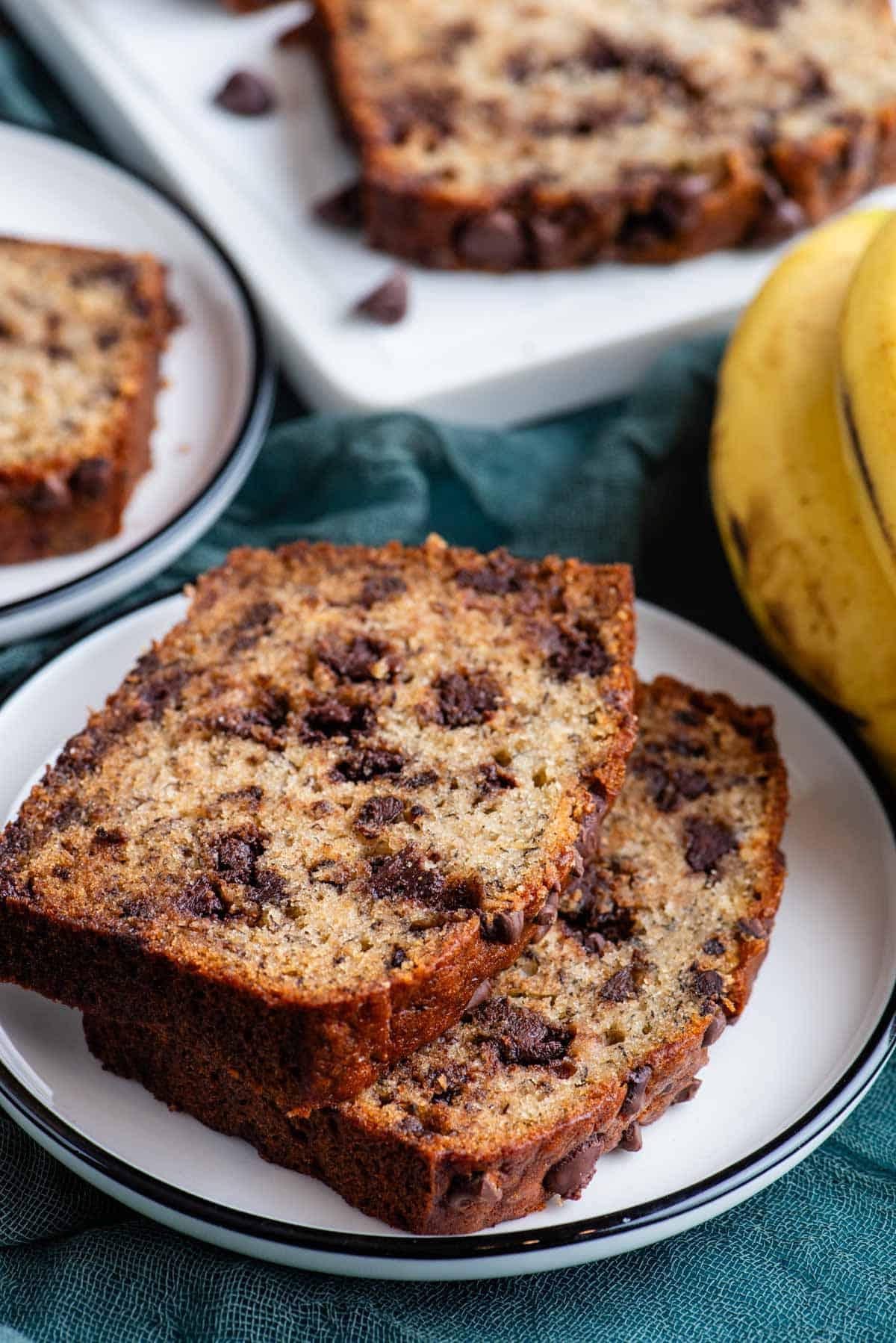 Due fette di pane alle banane e gocce di cioccolato su un piatto bianco sopra un asciugamano color ottanio scuro, con banane intere accanto, un altro piatto con una fetta di pane alle banane, e un tagliere bianco con altro pane dietro.