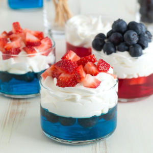 Tre bicchierini di vetro con bicchierini di gelatina festivi del 4 luglio: gelatina blu a strati con mirtilli e panna montata, uno sormontato da mirtilli extra, mentre gli altri hanno fragole su panna montata su gelatina rossa o blu.