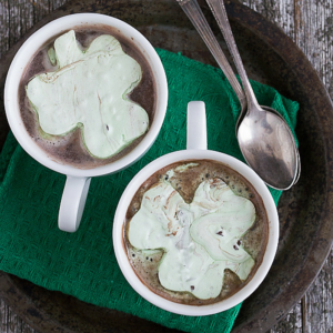 Due tazze di cioccolata calda con marshmallow verdi a forma di trifoglio su un vassoio scuro con due cucchiaini di metallo e un tovagliolo di stoffa verde, il tutto su una superficie di legno rustico.
