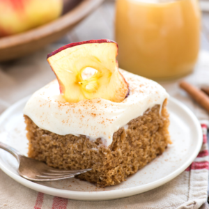 Una fetta di torta di mele speziata con glassa bianca, una spolverata di cannella e una fetta di mela essiccata, servita su un piatto bianco con una forchetta. Un bicchiere e della frutta sono sfocati sullo sfondo.