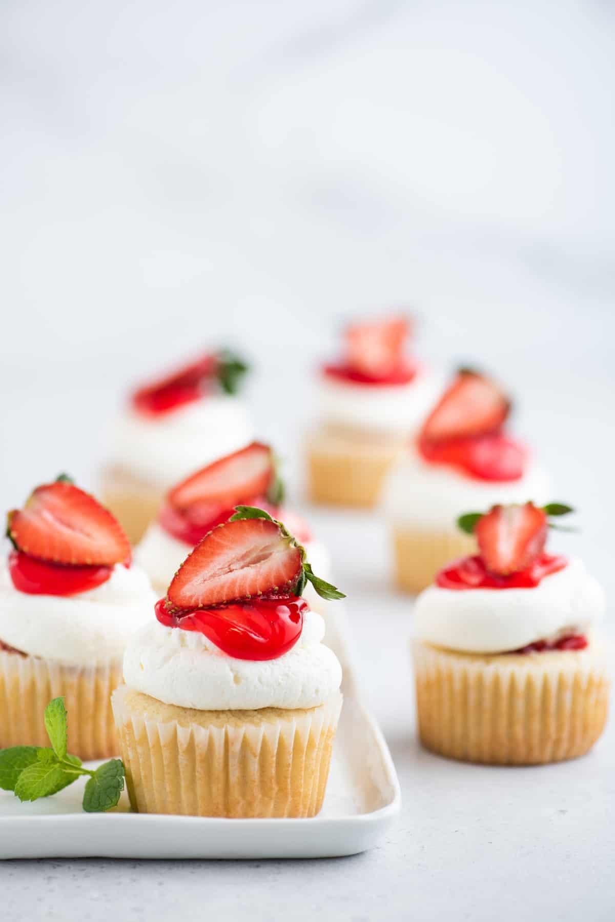 I Cupcake alle Fragole Shortcake sono fatti con torta alla vaniglia umida, ripieni di dolce salsa di fragole e ricoperti di soffice panna montata! Cupcake alle fragole shortcake su piatto bianco e tavolo