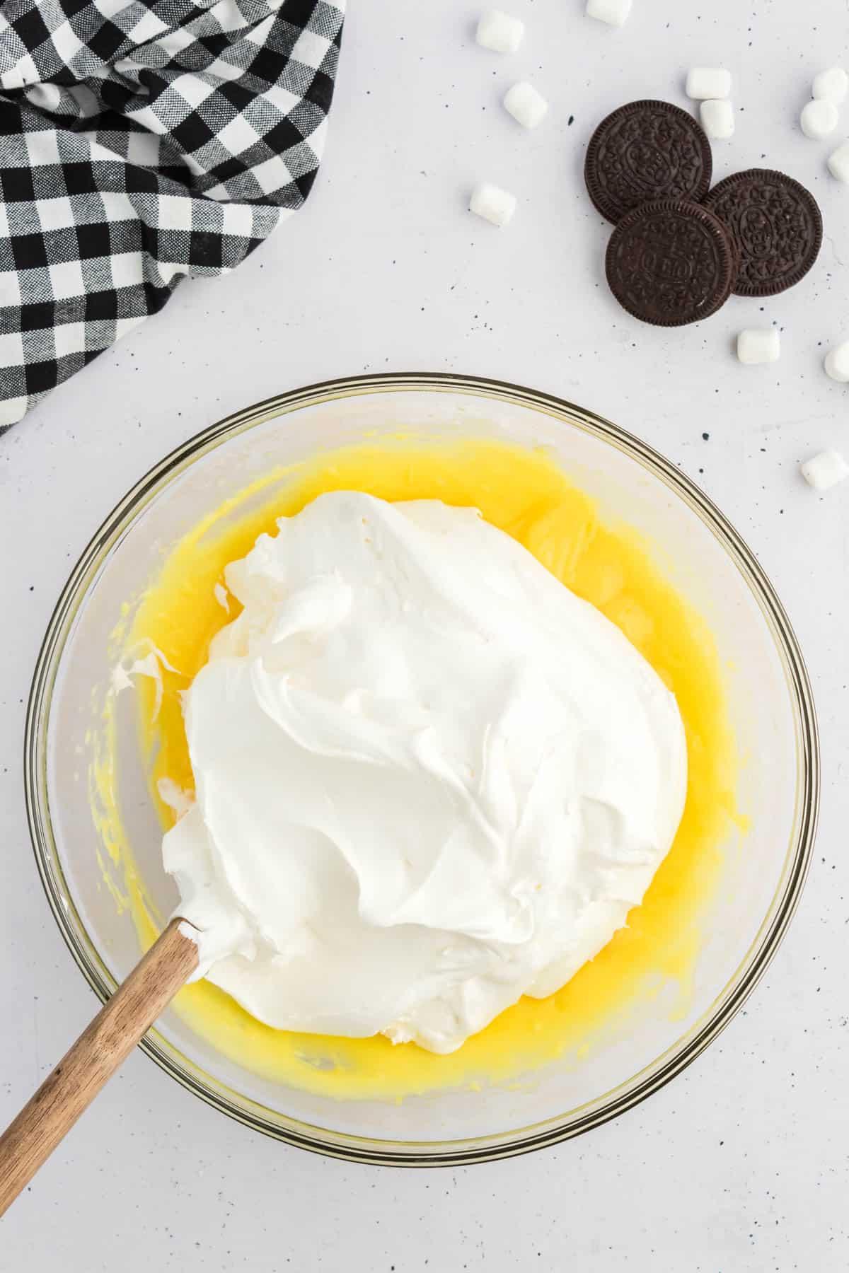 Budino alla vaniglia in una ciotola di vetro sormontato da Cool Whip, circondato da Oreo, mini marshmallow e un panno da cucina a scacchi bianchi e neri