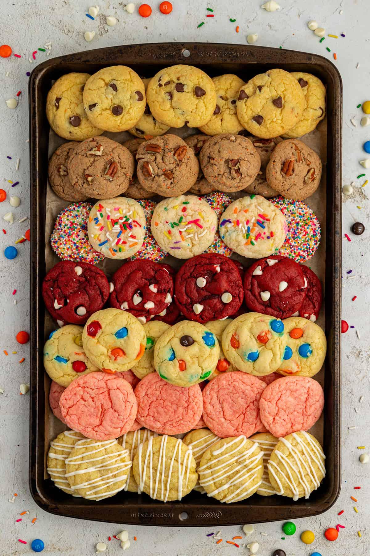 Fila di biscotti con preparato per torte di vari gusti impilati in diversi strati su una teglia circondata da M&M's, Reese's Pieces e codette colorate