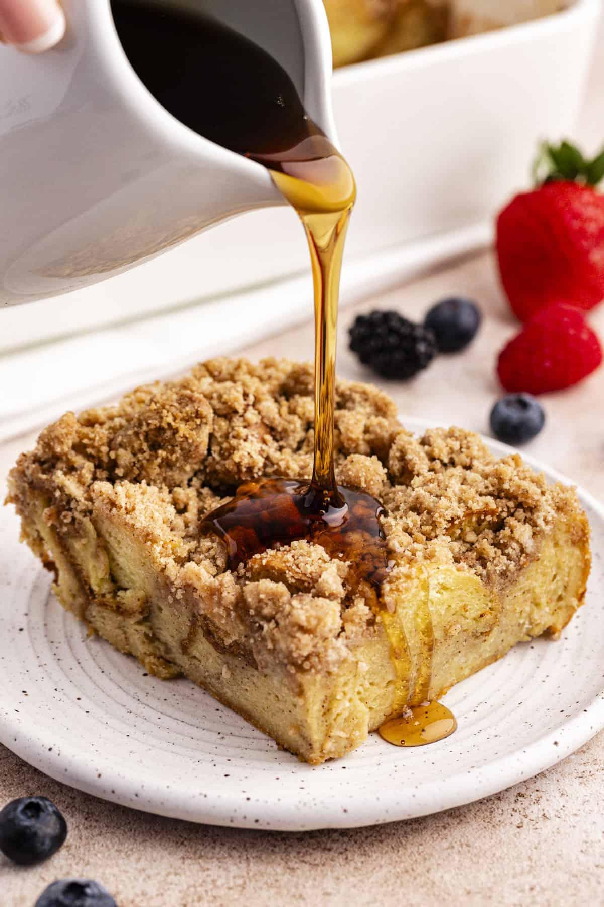 Una fetta di casseruola di French Toast su un piattino con sciroppo d'acero versato sopra e frutti di bosco freschi sparsi intorno.