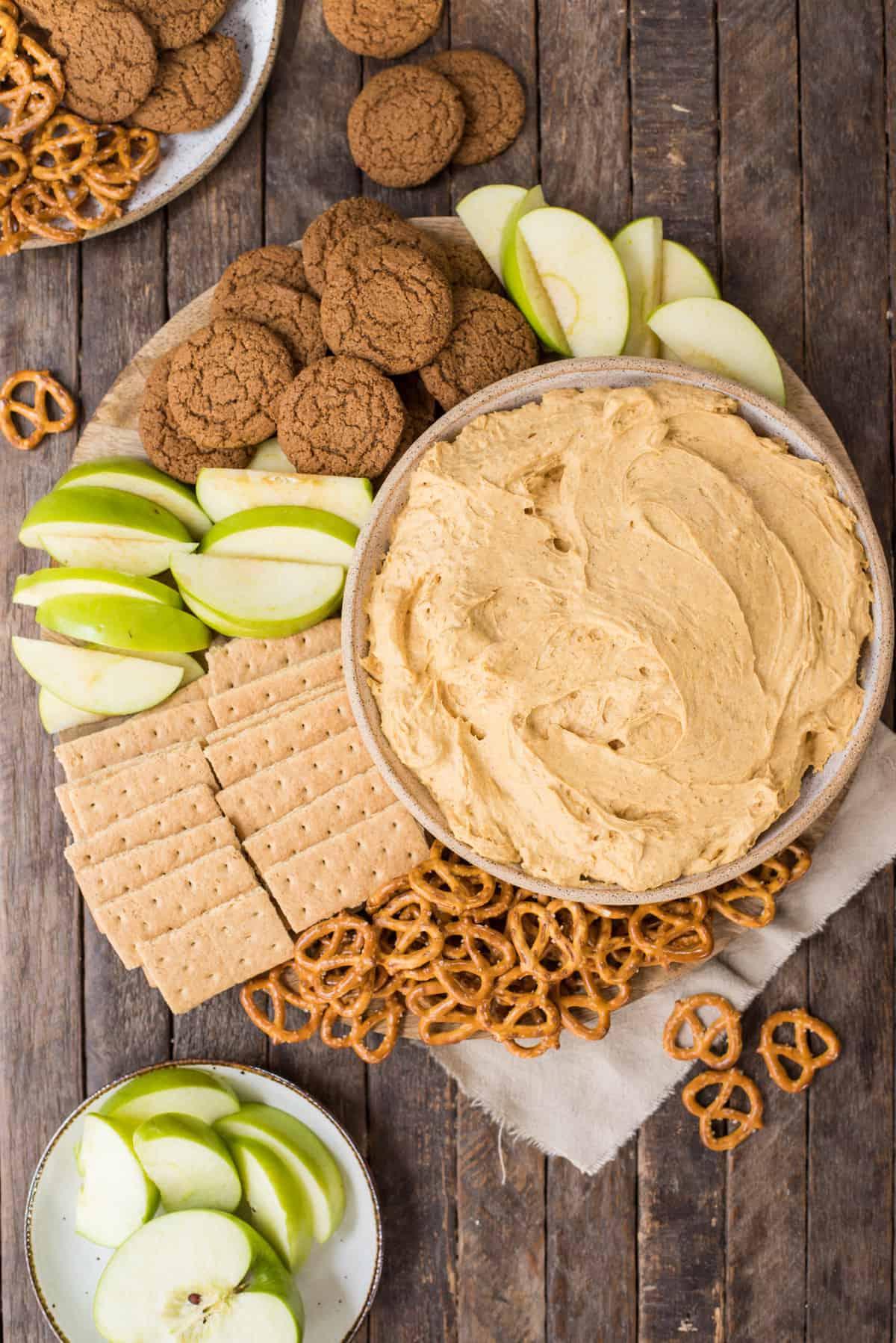 Cremosa crema di zucca in una ciotola circondata da pretzel, biscotti allo zenzero, cracker Graham e fette di mela verde su sfondo di legno.