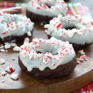 Ciambelle al Cioccolato e Bastoncino di Zucchero 6 Ciambelle al cioccolato guarnite con glassa bianca e caramelle alla menta piperita tritate, disposte su un tagliere di legno. Alcuni pezzi di ciambella al bastoncino di zucchero e pezzetti di menta sono sparsi intorno, creando un aspetto festivo.