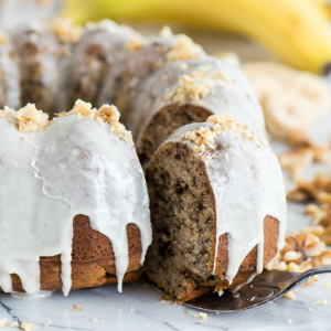 Una torta Bundt di banana e noci umida con glassa bianca che cola e noci tritate sopra; una fetta viene sollevata, rivelando la morbida consistenza della torta. Banane e altre noci sono sfocate sullo sfondo.