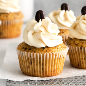 Primo piano di Muffin al Caffè con glassa cremosa a vortice e un chicco di caffè al cioccolato sopra, posizionati su carta forno e una griglia di raffreddamento.