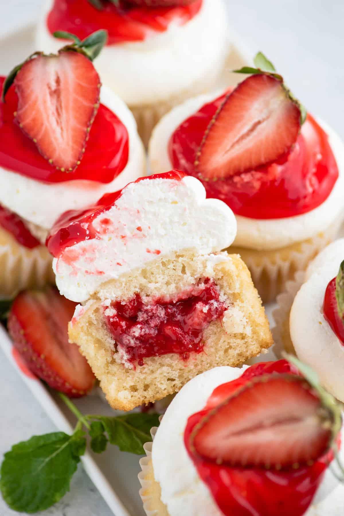 I Cupcake alle Fragole Shortcake sono fatti con torta alla vaniglia umida, ripieni di dolce salsa di fragole e ricoperti di soffice panna montata! Cupcake alle fragole shortcake, uno tagliato a metà per mostrare il ripieno