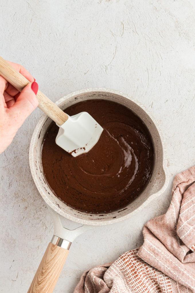 Ingredienti liquidi per la torta Texana in una casseruola mescolati con una spatola rossa e un panno da cucina color cuoio a sinistra