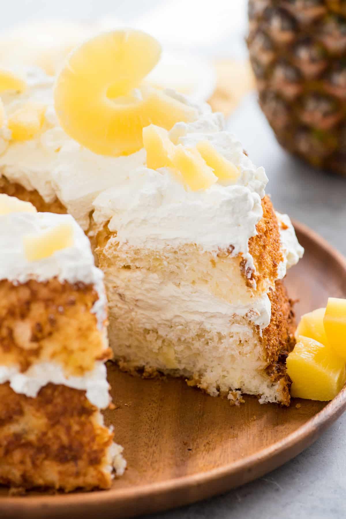 Torta degli angeli all'ananas 7 Torta Angel Food con fetta rimossa guarnita con panna montata e fette di ananas su vassoio di legno su sfondo bianco