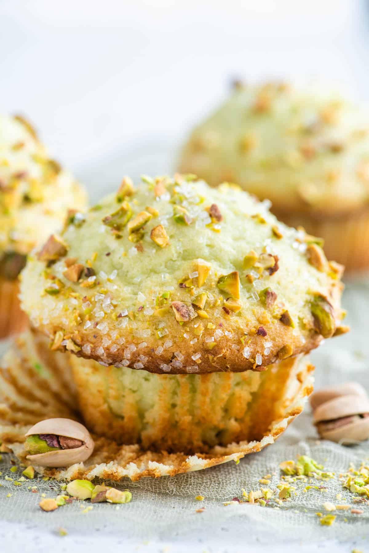 Muffin al Pistacchio 7 A pistachio muffin with its muffin paper pulled off, with whole pistachios around it and more muffins in the background.