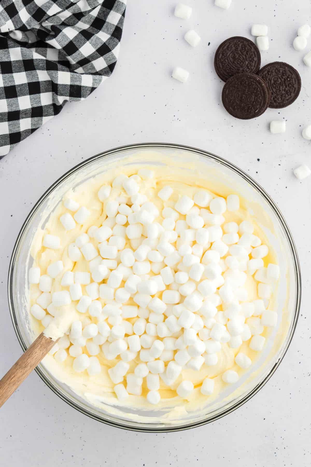 Miscela di budino alla vaniglia sormontata da mini marshmallow in una ciotola di vetro, circondata da Oreo, mini marshmallow e un panno da cucina a scacchi bianchi e neri
