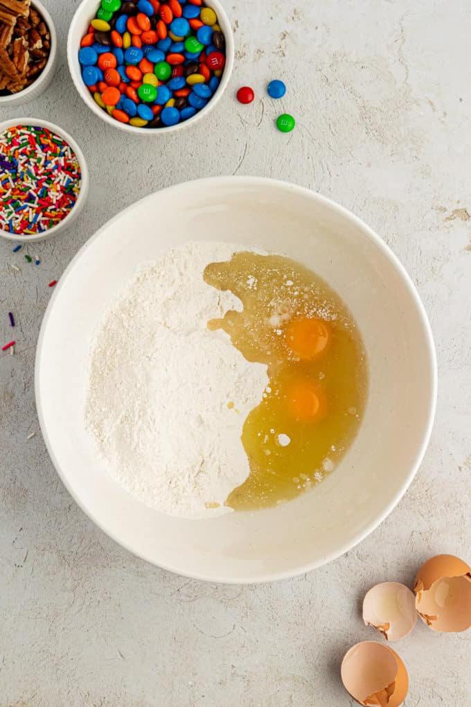 Uova e ingredienti secchi per biscotti con preparato per torte in una ciotola bianca circondata da gusci d'uovo, una ciotola di codette, una ciotola di M&M's e una ciotola di noci