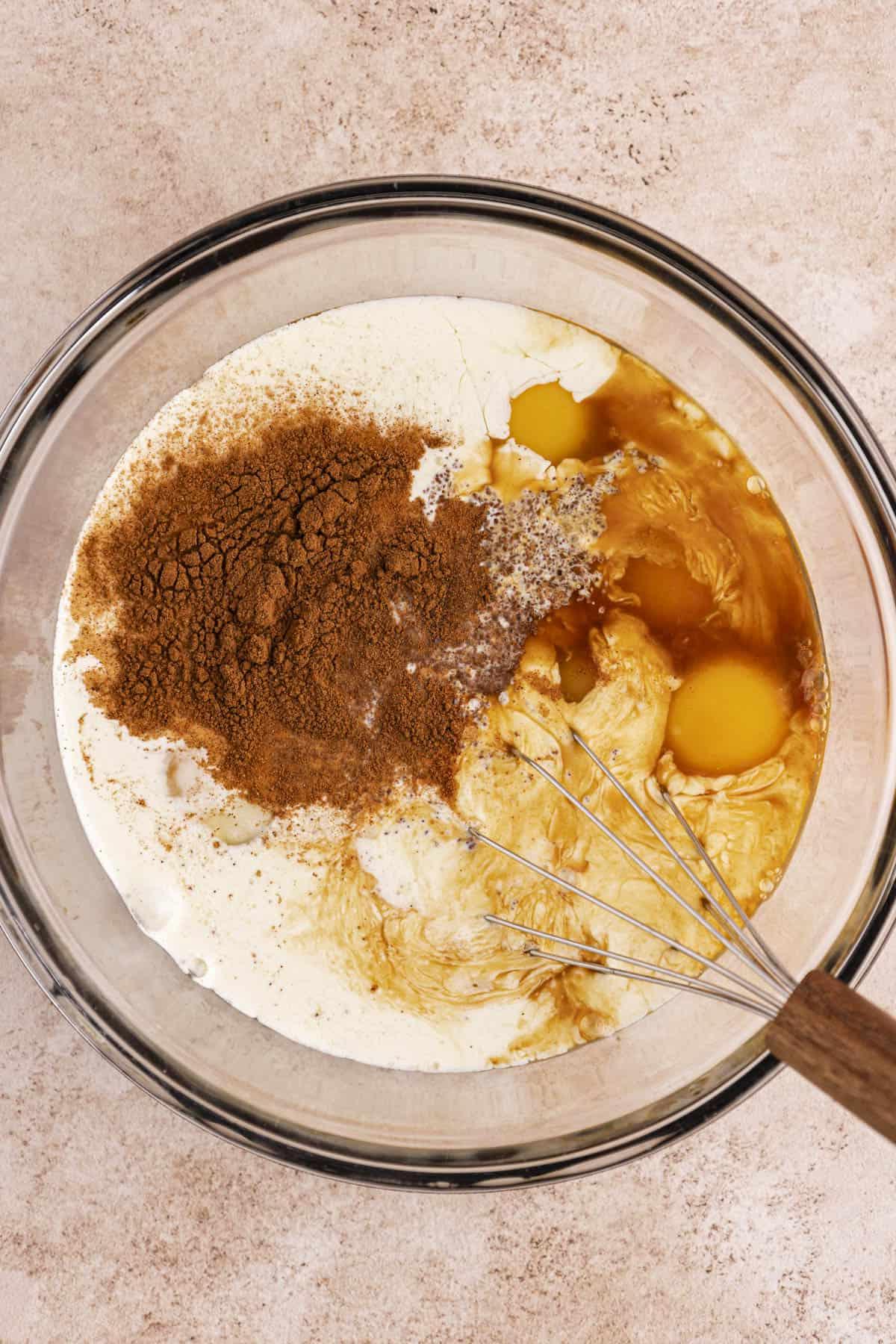 Una ciotola di vetro trasparente con uova, latte intero, panna fresca, zucchero di canna, cannella, sale ed estratto di vaniglia con una frusta appoggiata all'interno della ciotola.