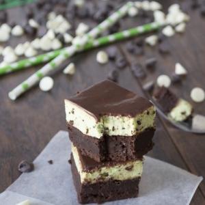 Tre brownies al cioccolato impilati con uno strato cremoso di menta al centro sono posti su un pezzo di carta forno. Gocce di cioccolato bianco e fondente e cannucce verdi sono sparse su una superficie di legno sullo sfondo.
