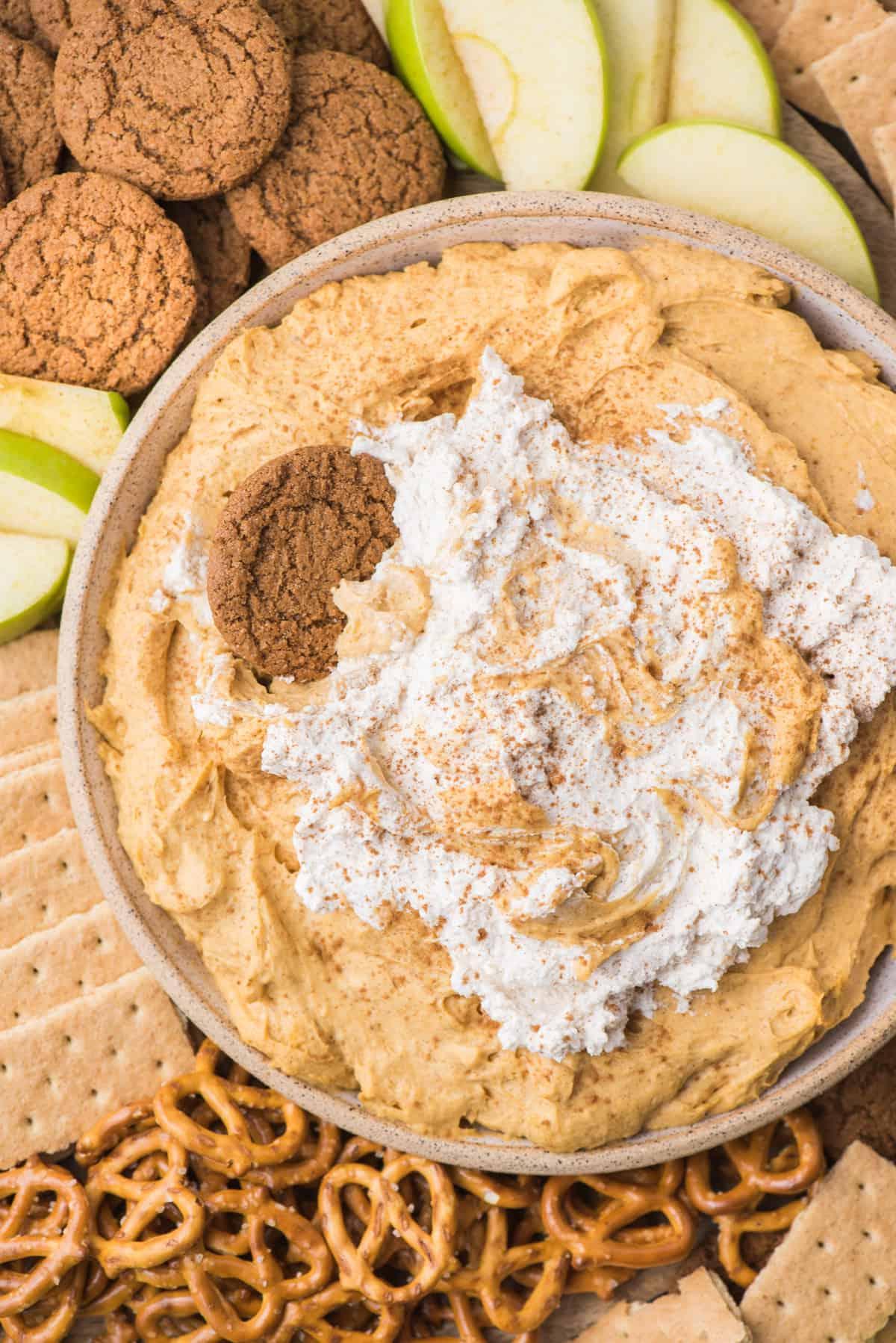 Cremosa crema di zucca con panna montata in una ciotola, circondata da pretzel, biscotti allo zenzero, cracker Graham e fette di mela verde su un piatto di legno.